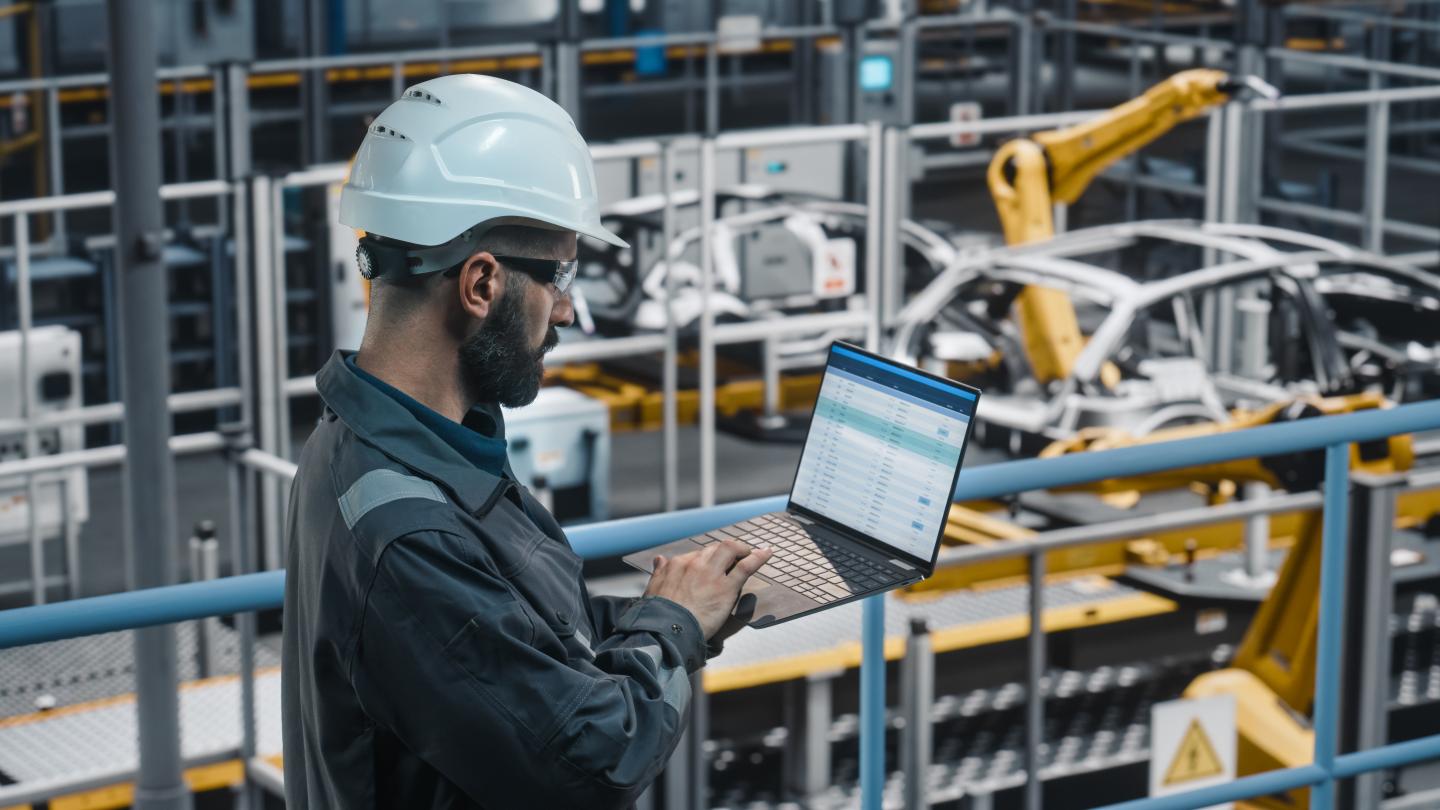 Car Factory Engineer in Work Uniform Using Laptop Computer with Spreadsheet Software.