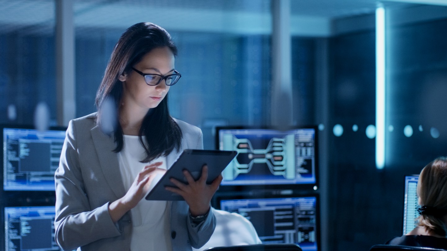 Young Female Employee Wearing Glasses Uses Tablet in System Control Center. In the Background Her Coworkers are at Their Workspaces with many Displays Showing Valuable Data.