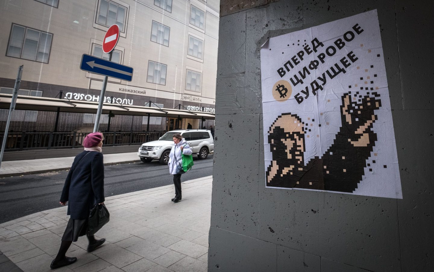 Russian women walk past a poster of Lenin and a bitcoin.