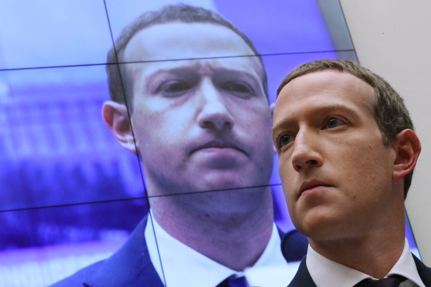 Mark Zuckerberg looks off camera while standing in front of a jumbo screen displaying his own face, staring down at him