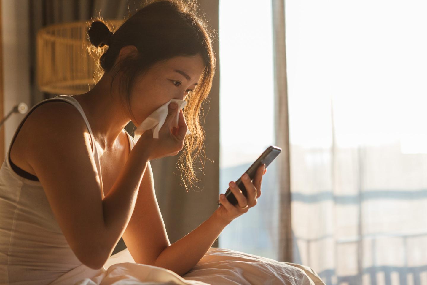Young woman sneezing while using mobile phone in bed