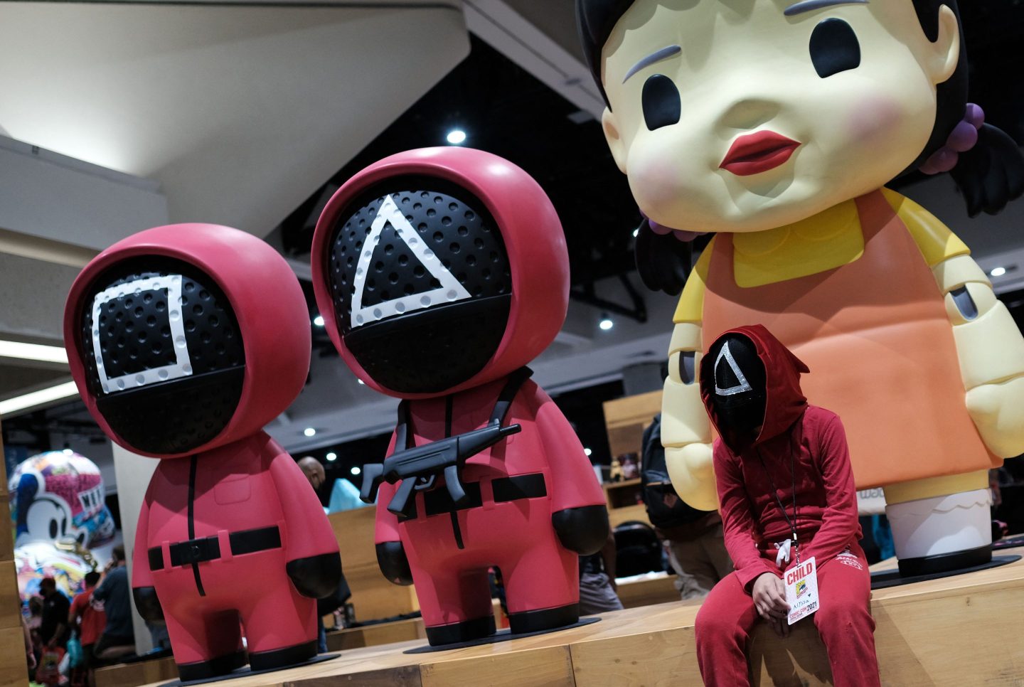 A child in a pink Squid Game costume sits next to three life size, cartoonish Squid Game figurines at San Diego Comic Con in 2021