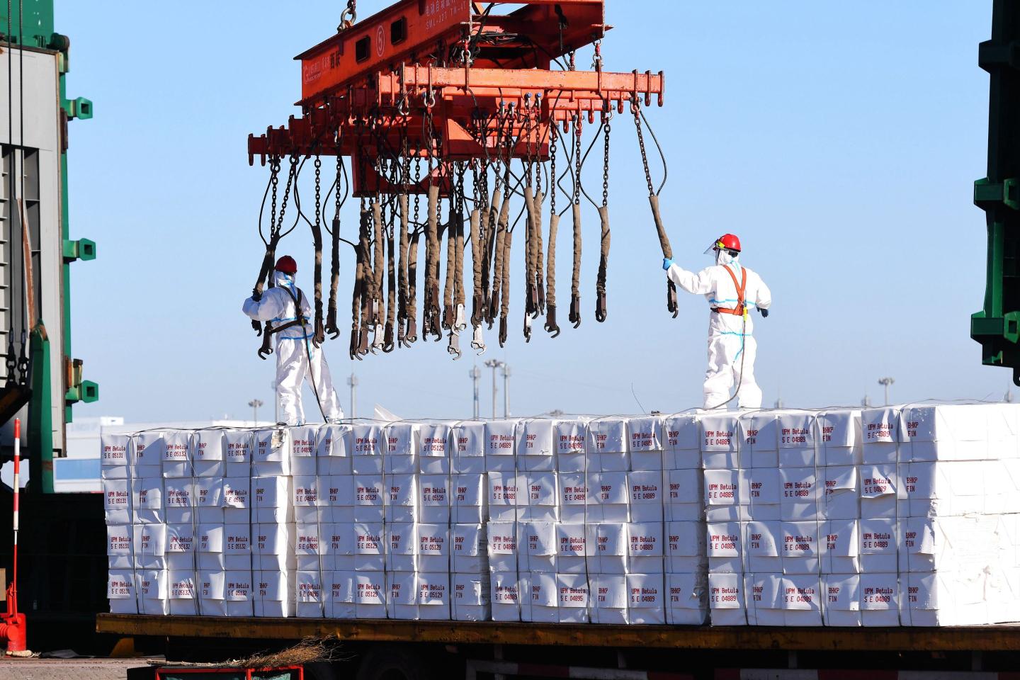 Two port workers in personal protective gear help load goods onto a ship