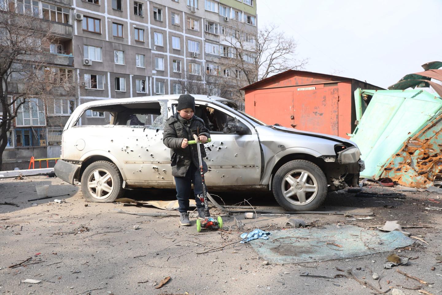 a young boy stands in front of car damaged by war.