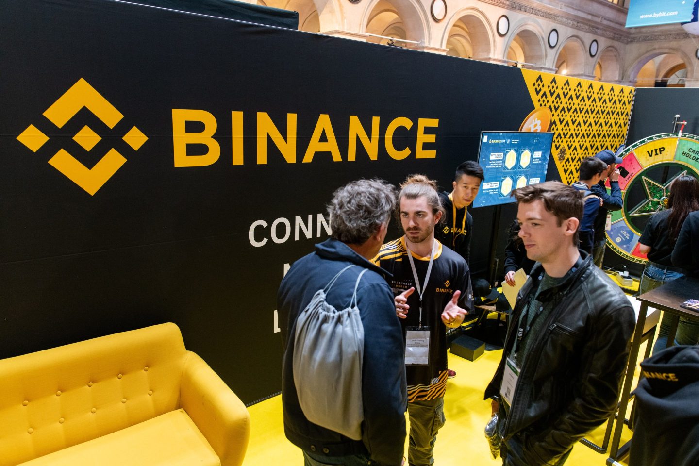 The Binance logo is seen behind people in conversation at a Binance booth during a crypto event