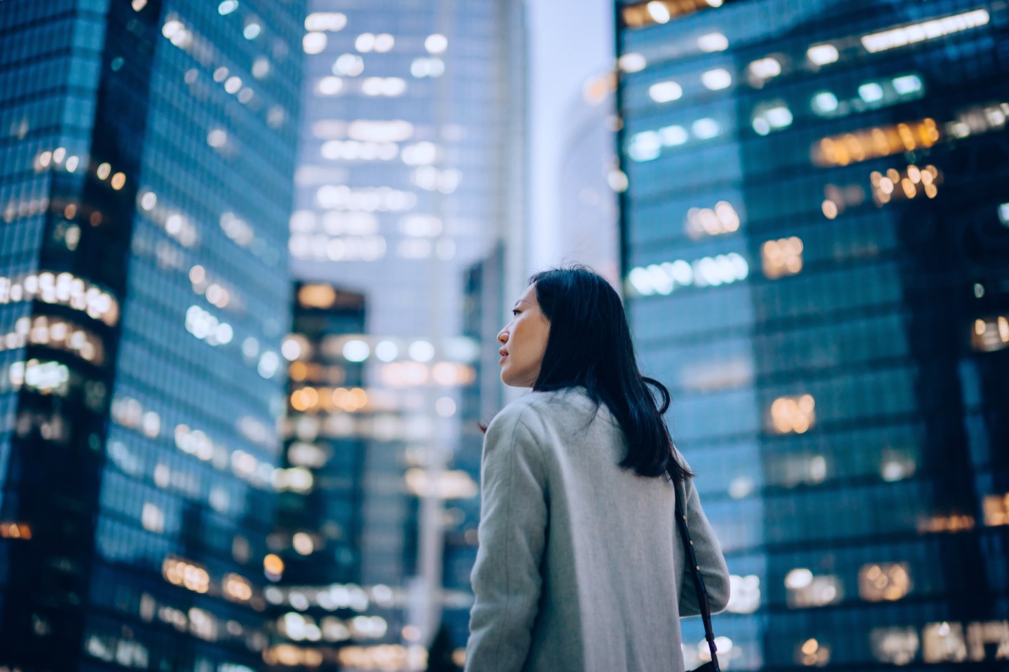Low angle side profile of confident and professional young Asian businesswoman looking up while standing against contemporary corporate skyscrapers with illuminated facade in financial district in the evening.