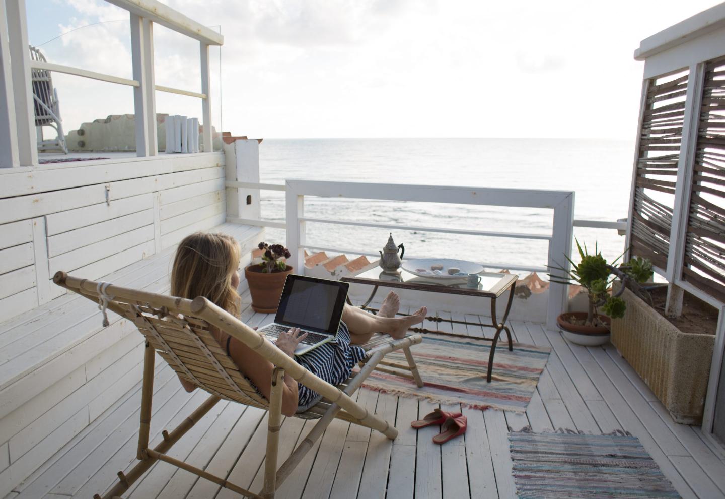 A woman works on a laptop from a seaside deck