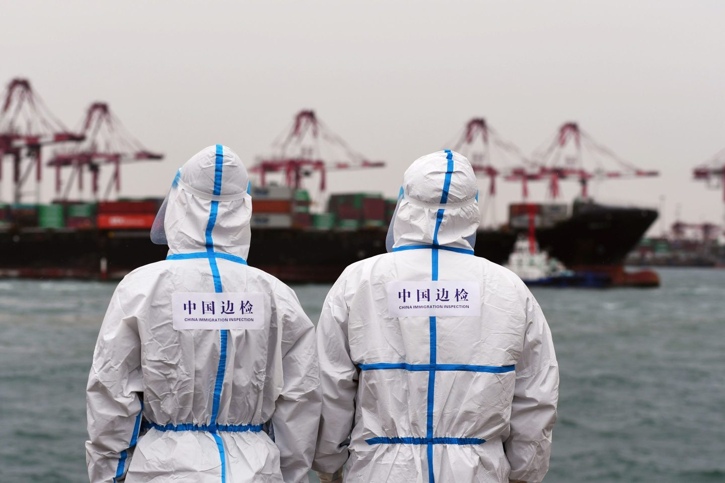 Two health workers in hazmat suits look across the water to a ship berthed in the Chinese port of Qingdao