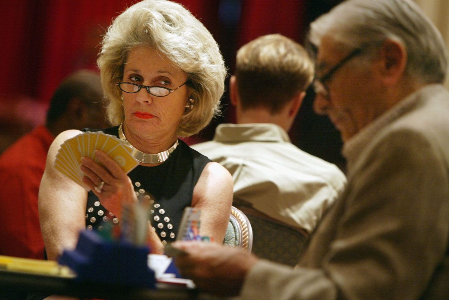 A woman with big hair arches her eyeborws and peers over her cards at an opposing player during a bridge tournament.