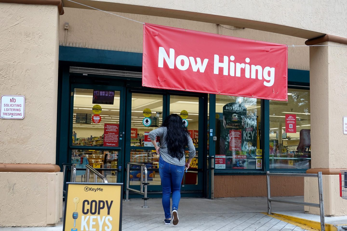 A woman walks into a store with a large now hiring sign hanging above the entrance.