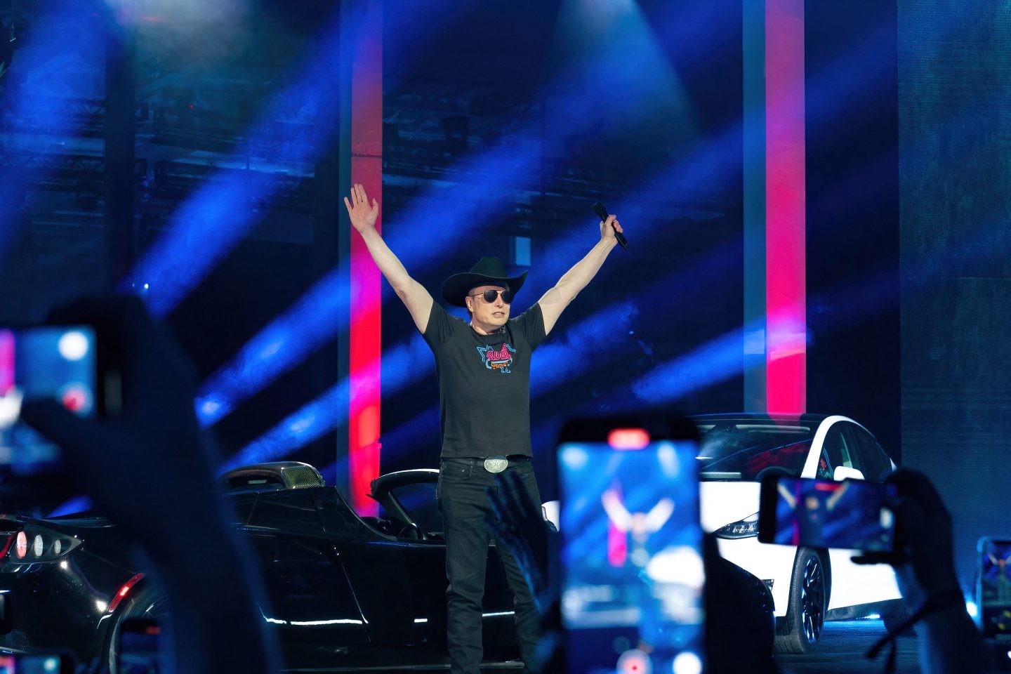 Elon Musk on stage at Tesla Giga Texas event with hands raised.