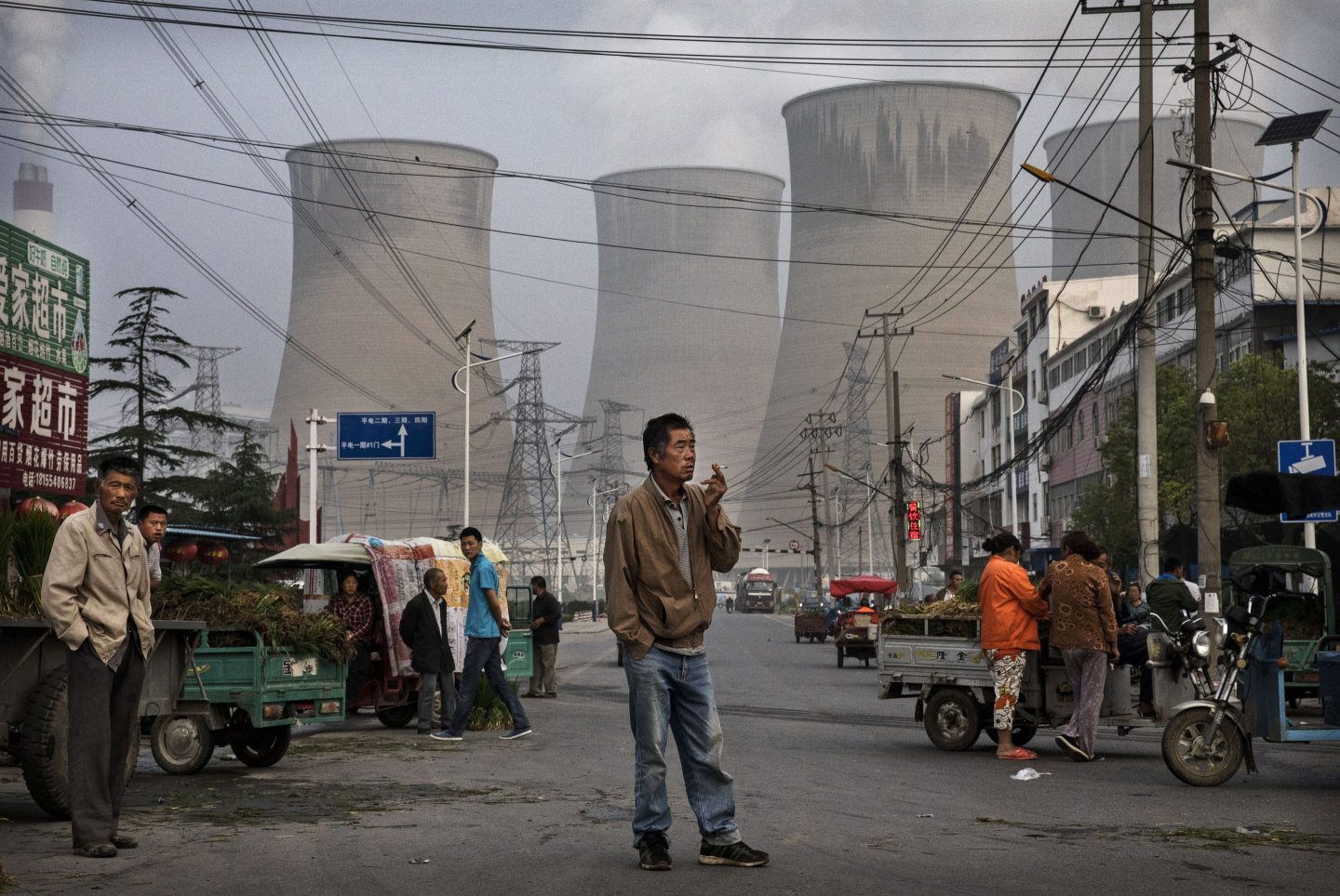 A man stands smoking a cigarette with coal power plants behind him.