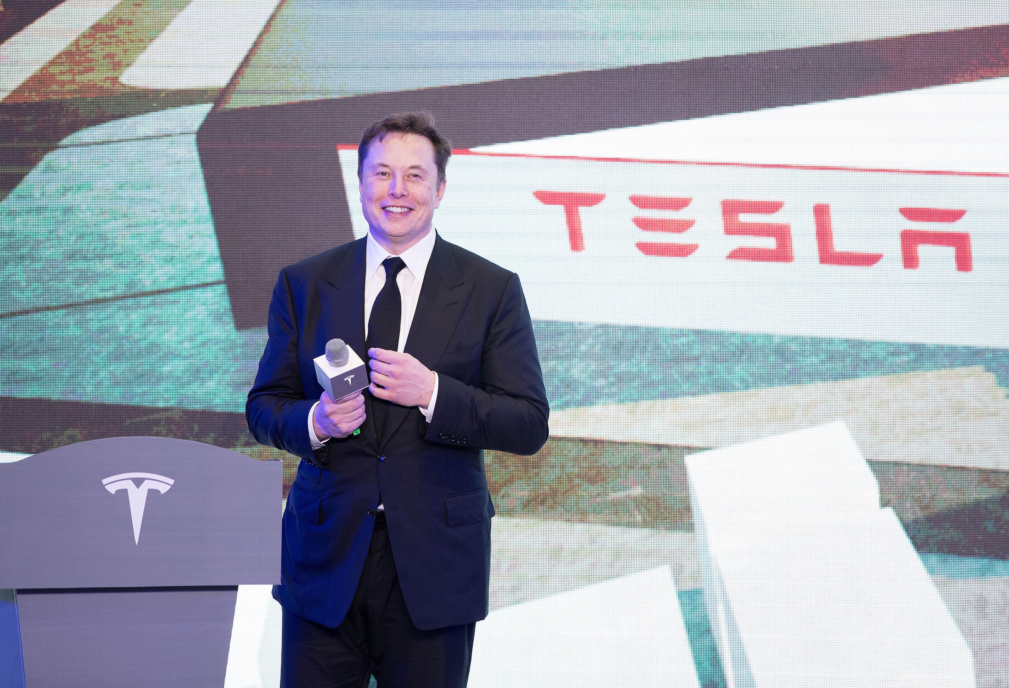 Elon Musk is seen onstage in front of a screen displaying the Tesla logo.