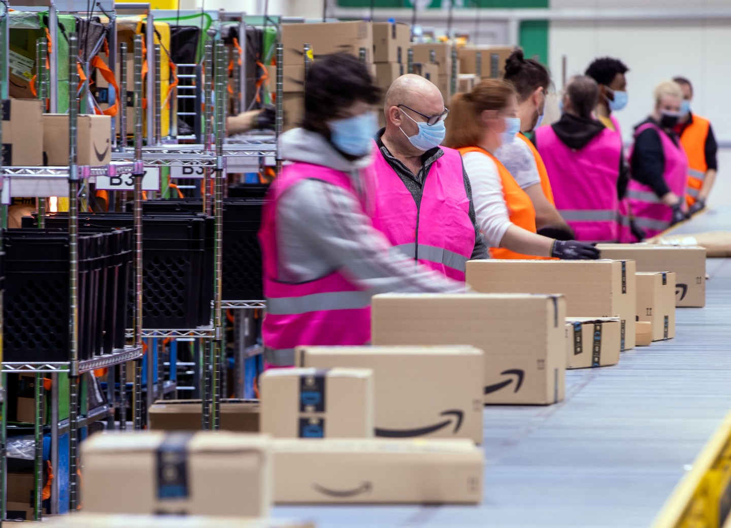 Several workers, wearing high-vis vests, are seen sorting packages along a conveyer belt at an Amazon warehouse.