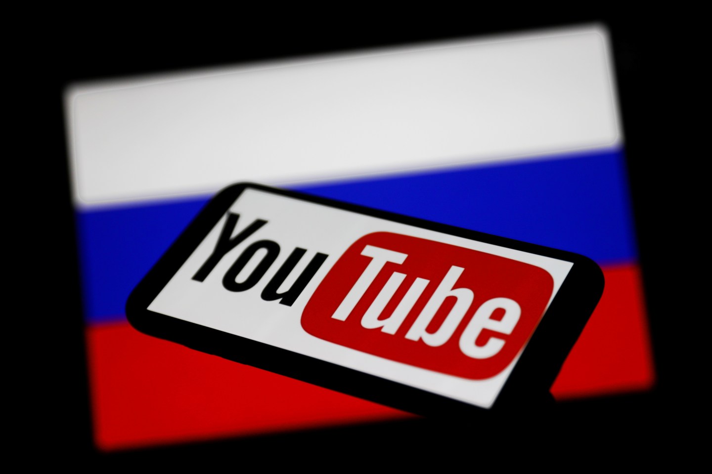 YouTube logo displayed on a phone screen and Russian flag displayed on a screen in the background are seen in this illustration photo taken in Krakow, Poland on March 1, 2022.