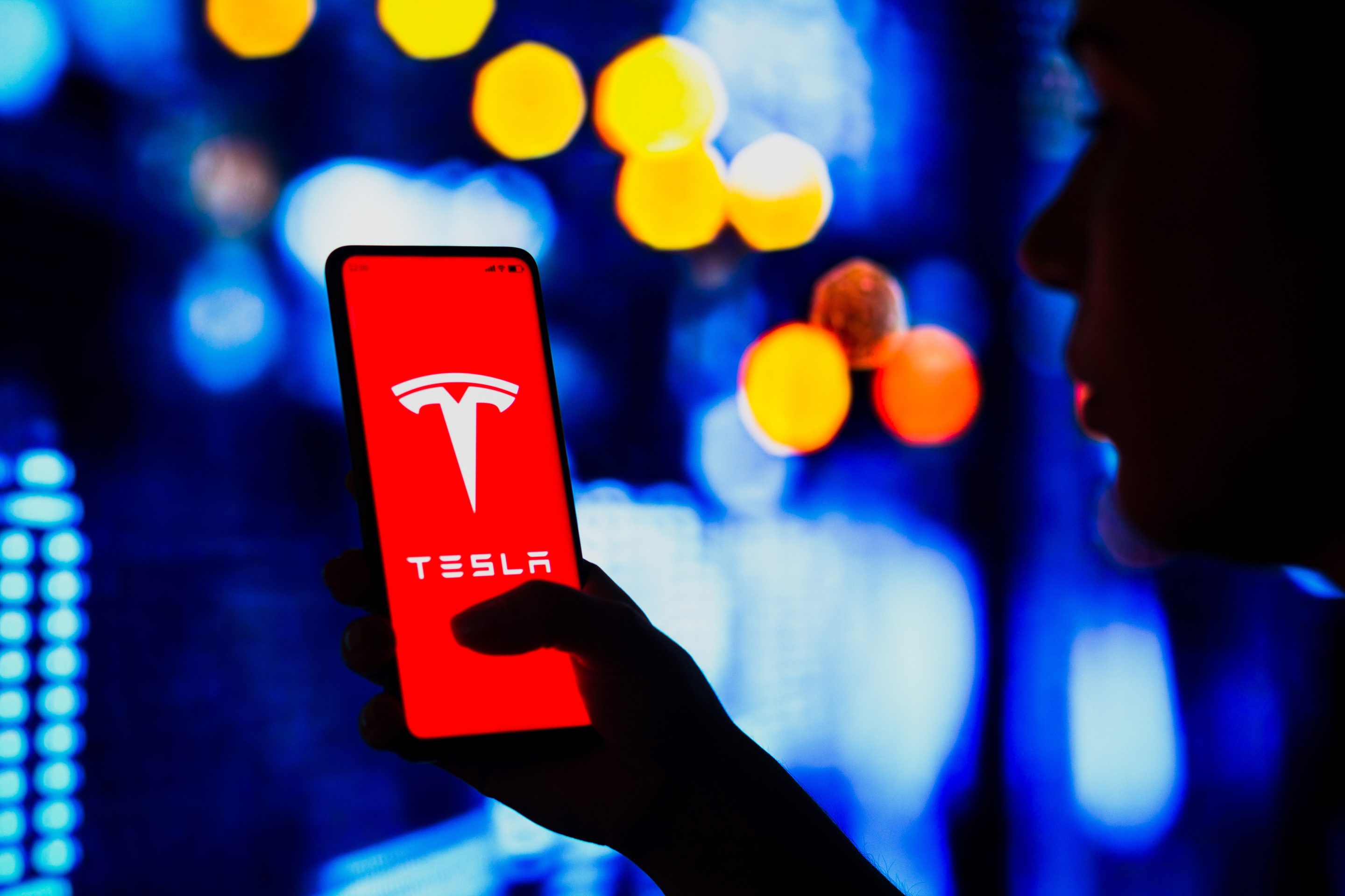 a silhouetted woman holds a smartphone with the Tesla logo displayed on the screen