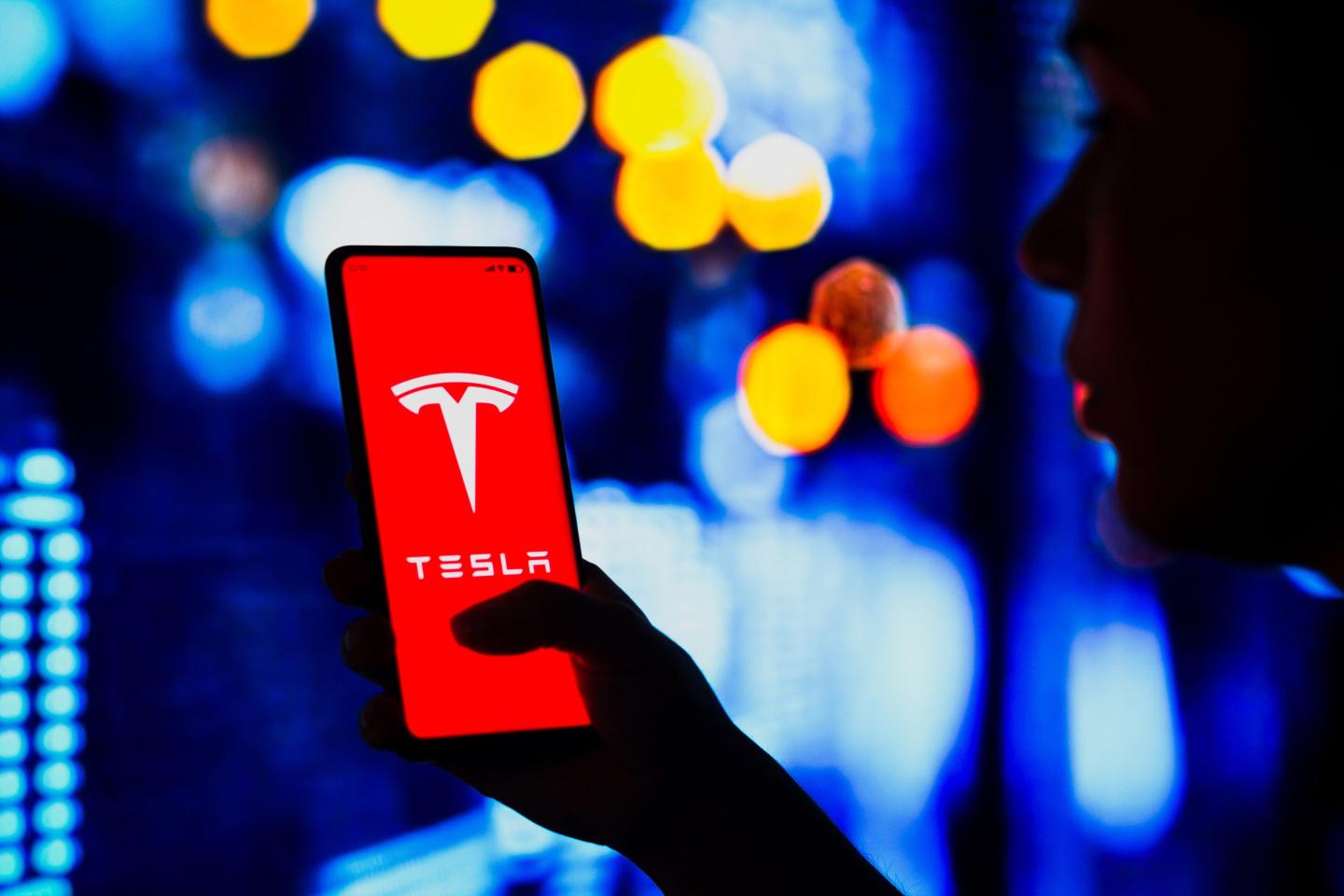 a silhouetted woman holds a smartphone with the Tesla logo displayed on the screen