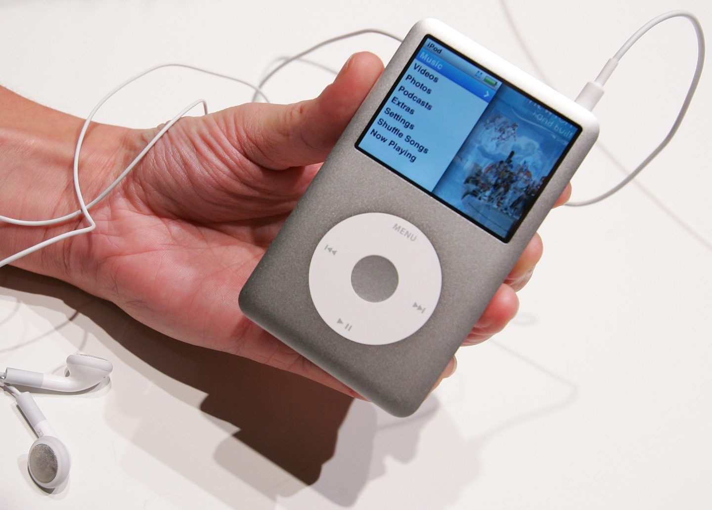 An iPod Classic is held by an unseen person. The device's screen displays the main menu.