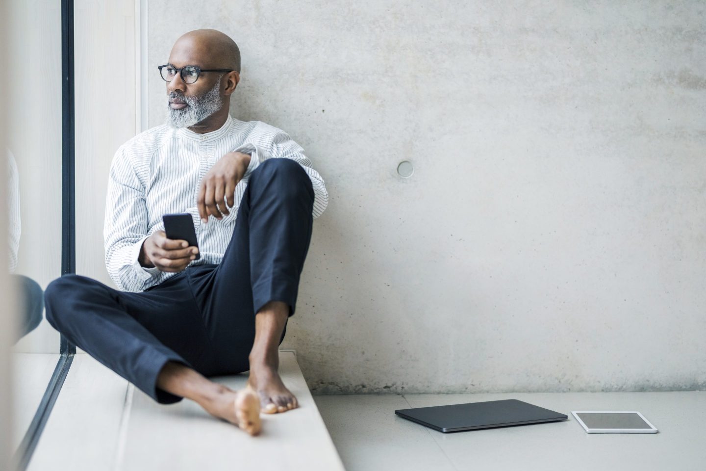 Barefoot mature businessman with smartphone sitting on window sill looking at distance