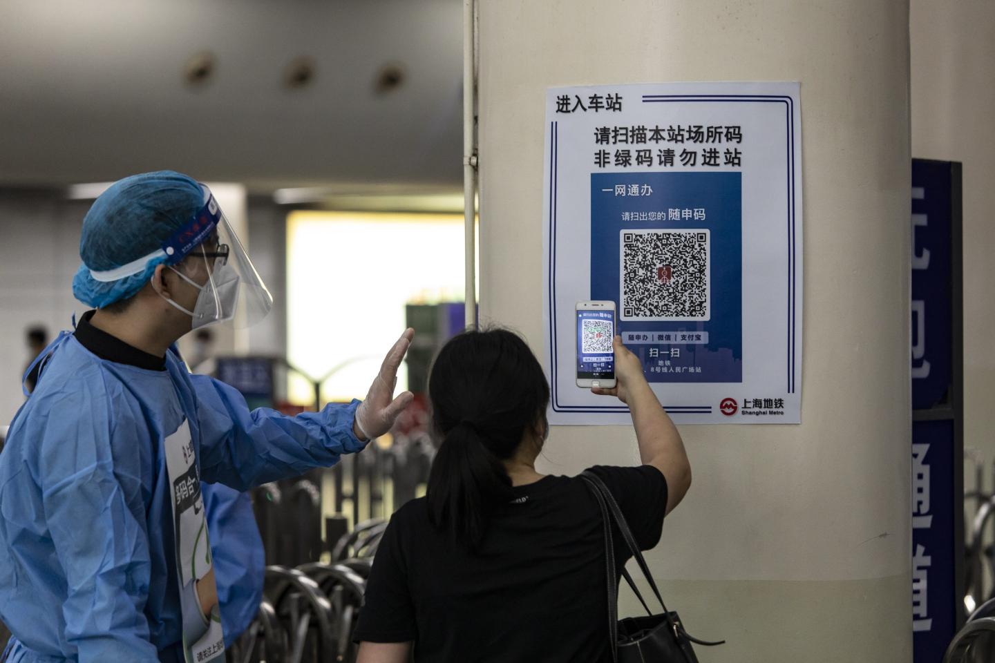 A man in PPE assists a woman as she holds up her smartphone to scan a QR code printed on a poster next to a subway entrance in China