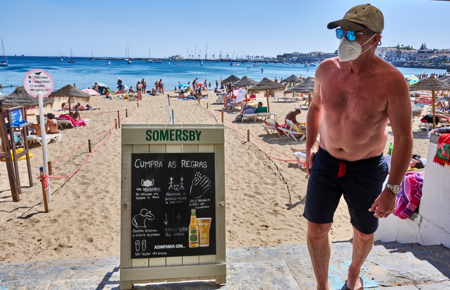 CASCAIS, PORTUGAL &#8211; AUGUST 18: A mask-clad beachgoer walks by a COVID-19 warning sign on the promenade above Praia da Duquesa on a sunny and warm Summer day during the coronavirus pandemic on August 18, 2021 in Cascais, Portugal. Although restrictions are being lifted in steps from August 01 tourism and everyday activities are yet to reach pre pandemic levels in Portugal, where 17 more deaths and 2,983 new cases related to COVID-19 were recorded in the last 24 hours.  (Photo by Horacio Villalobos#Corbis/Corbis via Getty Images)