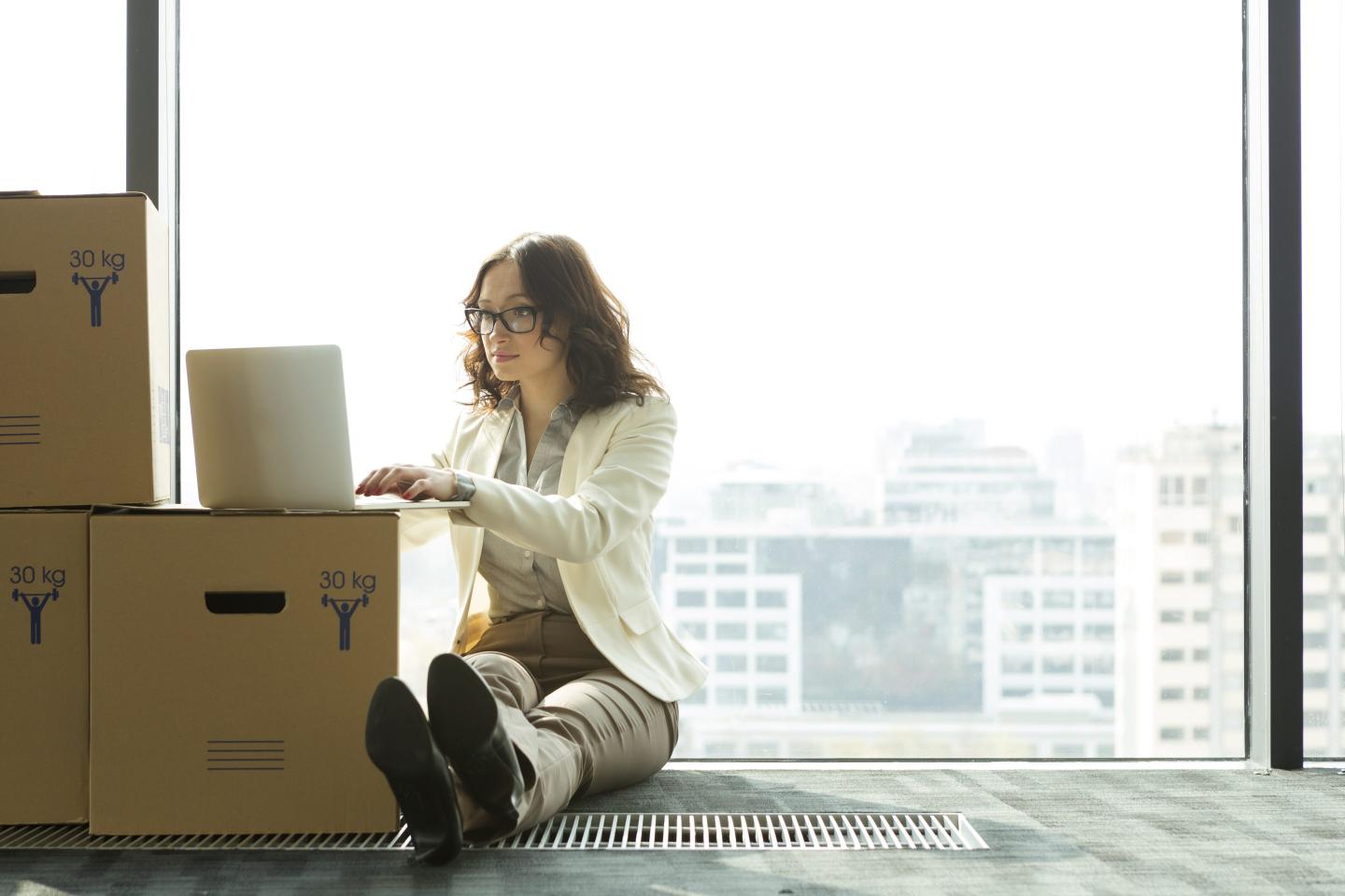 Businesswoman using laptop on empty office floor with cardboard boxes
