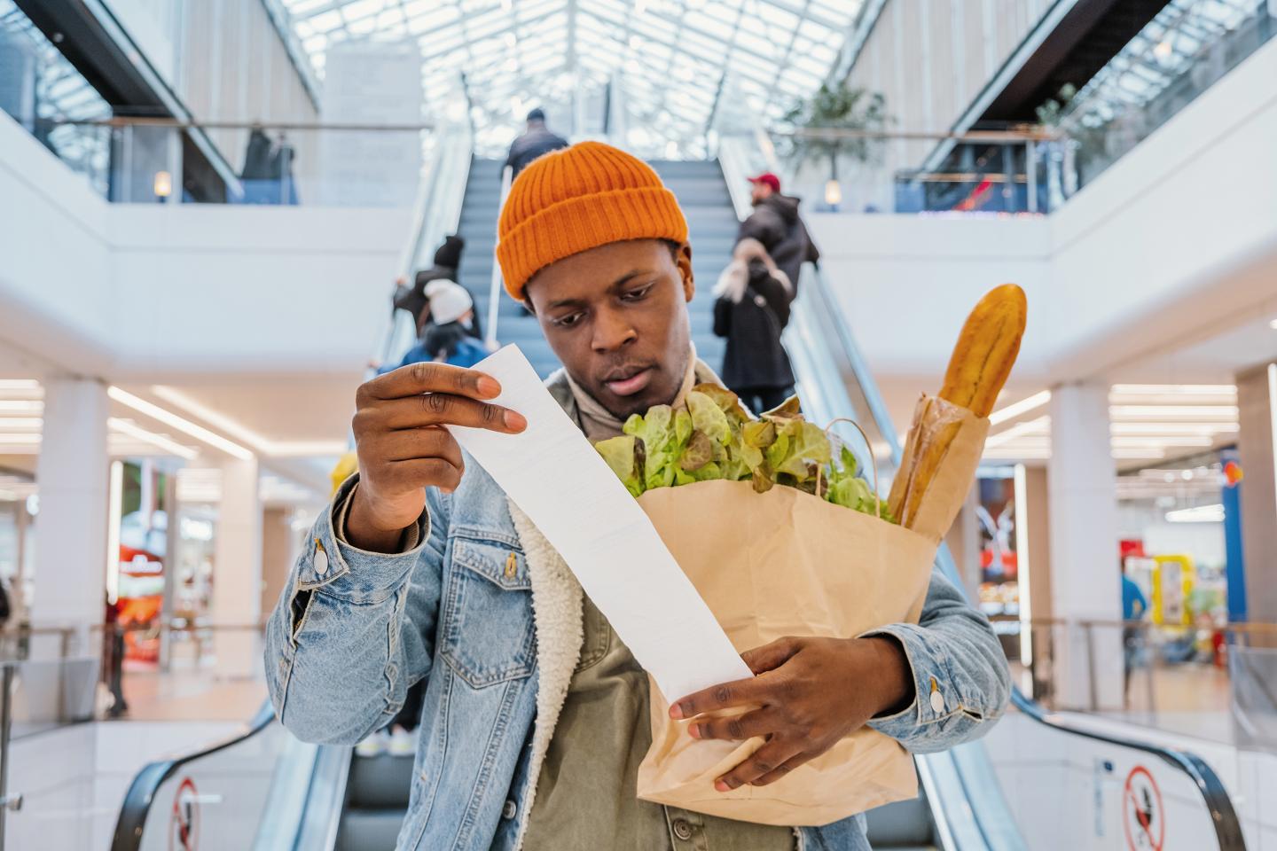 Surprised man looks at receipt total in sales check holding paper bag with groceries.