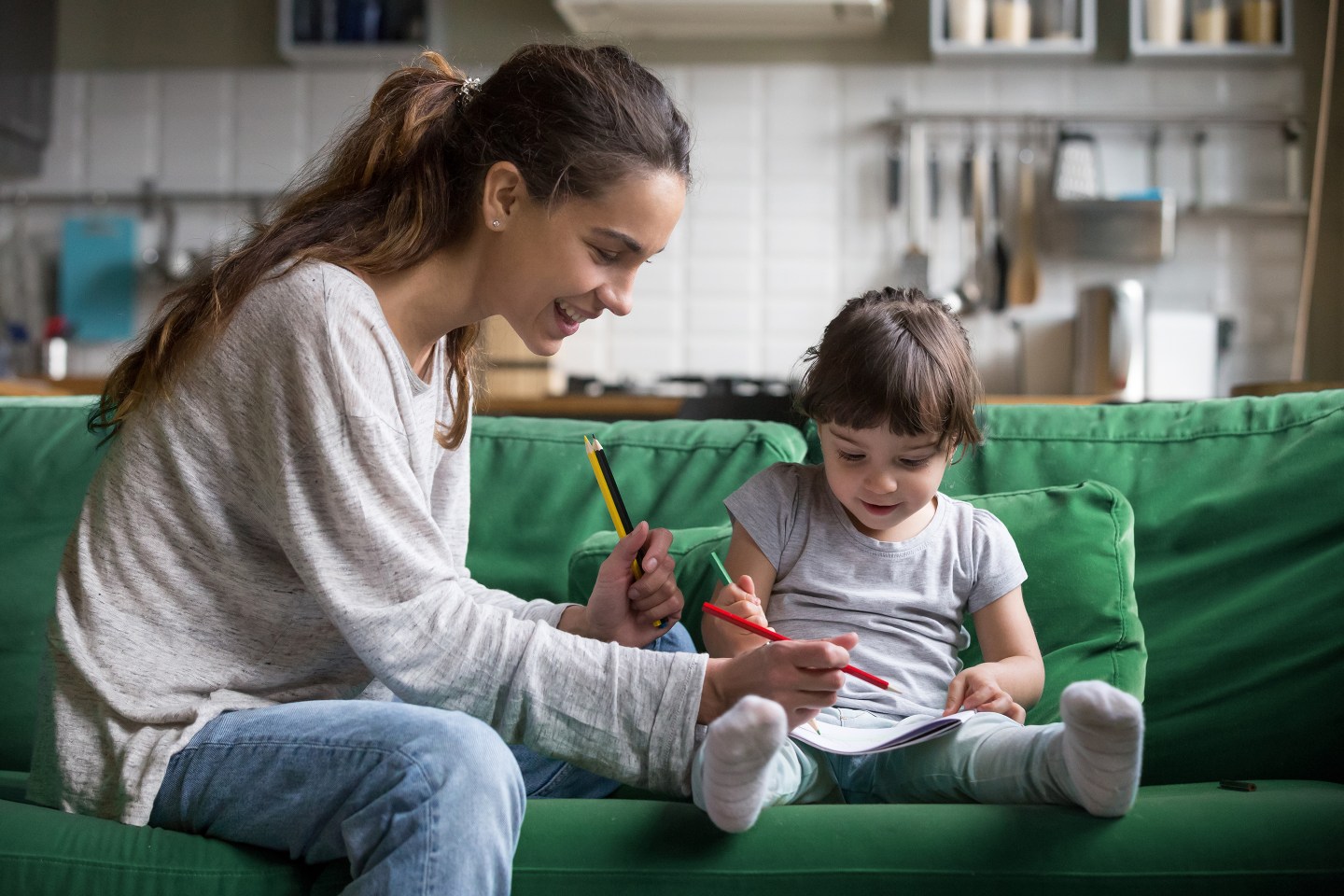Smiling baby sitter and preschool kid girl drawing with colored pencils sitting on sofa together.