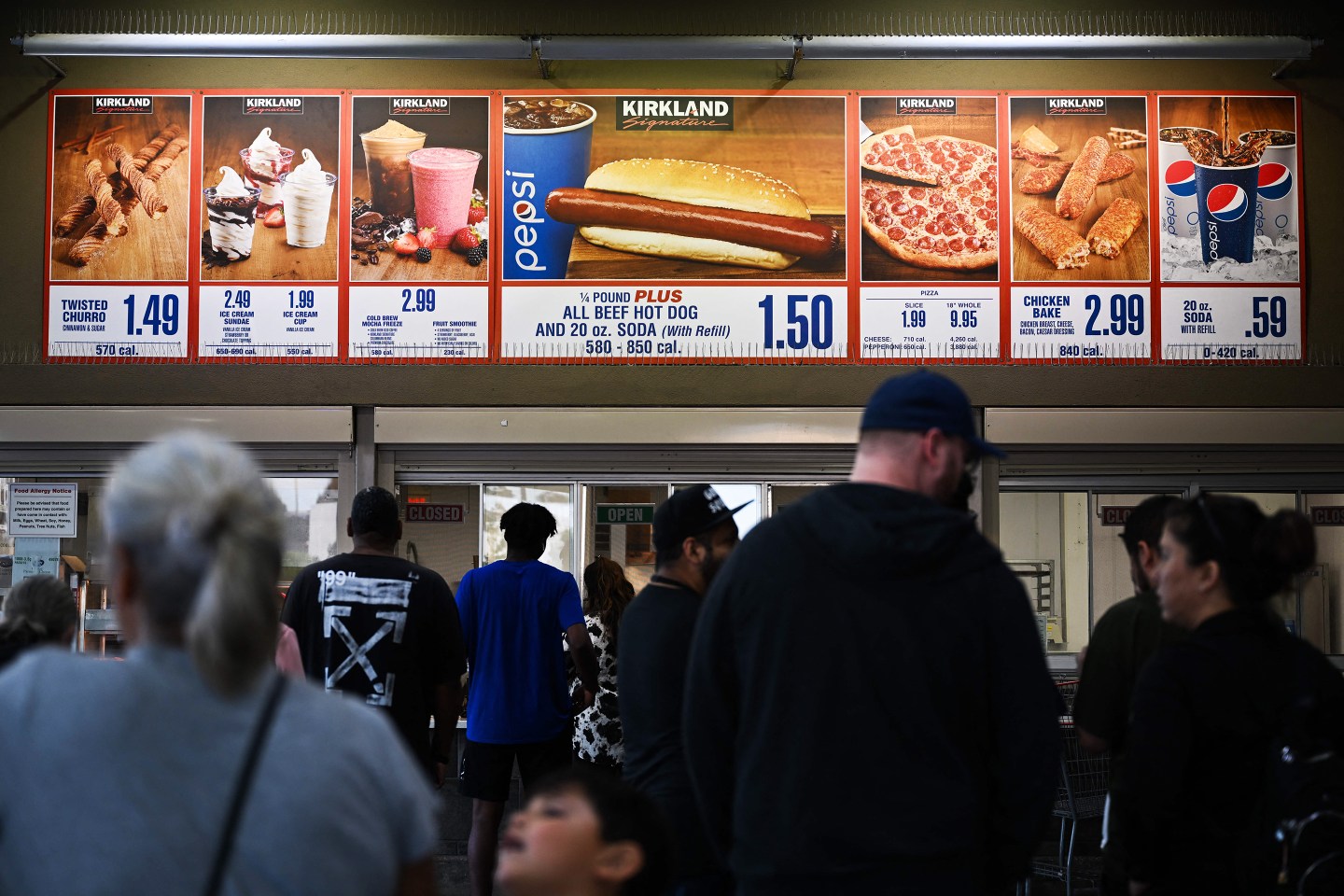 Customers wait in line to order below signage for the Costco Kirkland Signature $1.50 hot dog and soda combo, which has maintained the same price since 1985 despite consumer price increases and inflation.