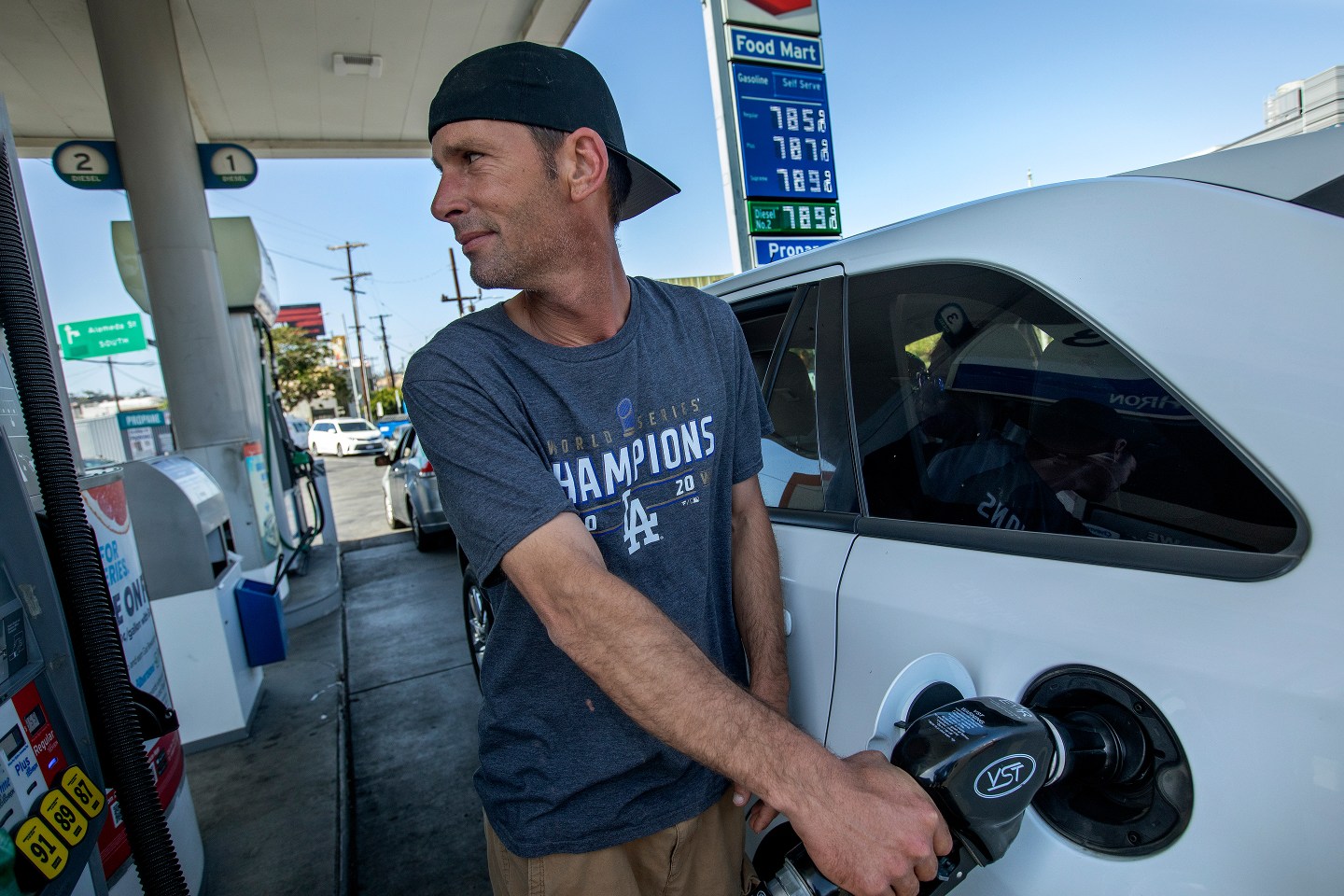 A man fills up his car with gas with a sign in the background that says regular gas is $7.85.