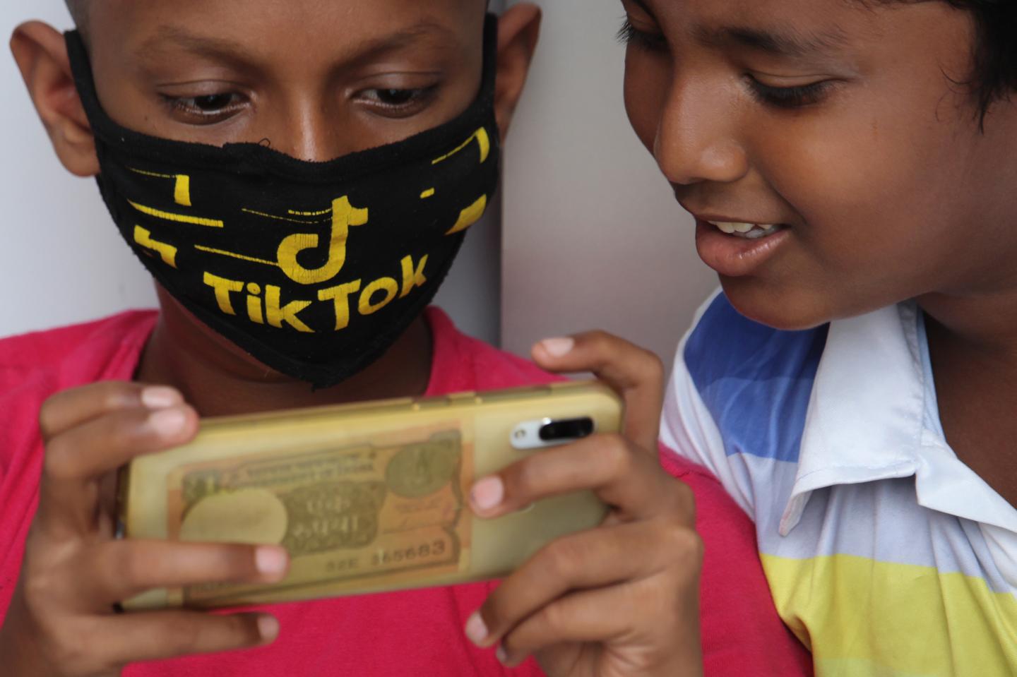 A boy wearing a face mask with the TikTok logo uses a mobile phone outside the downed shutters of a shop in Mumbai, India on July 29, 2020.