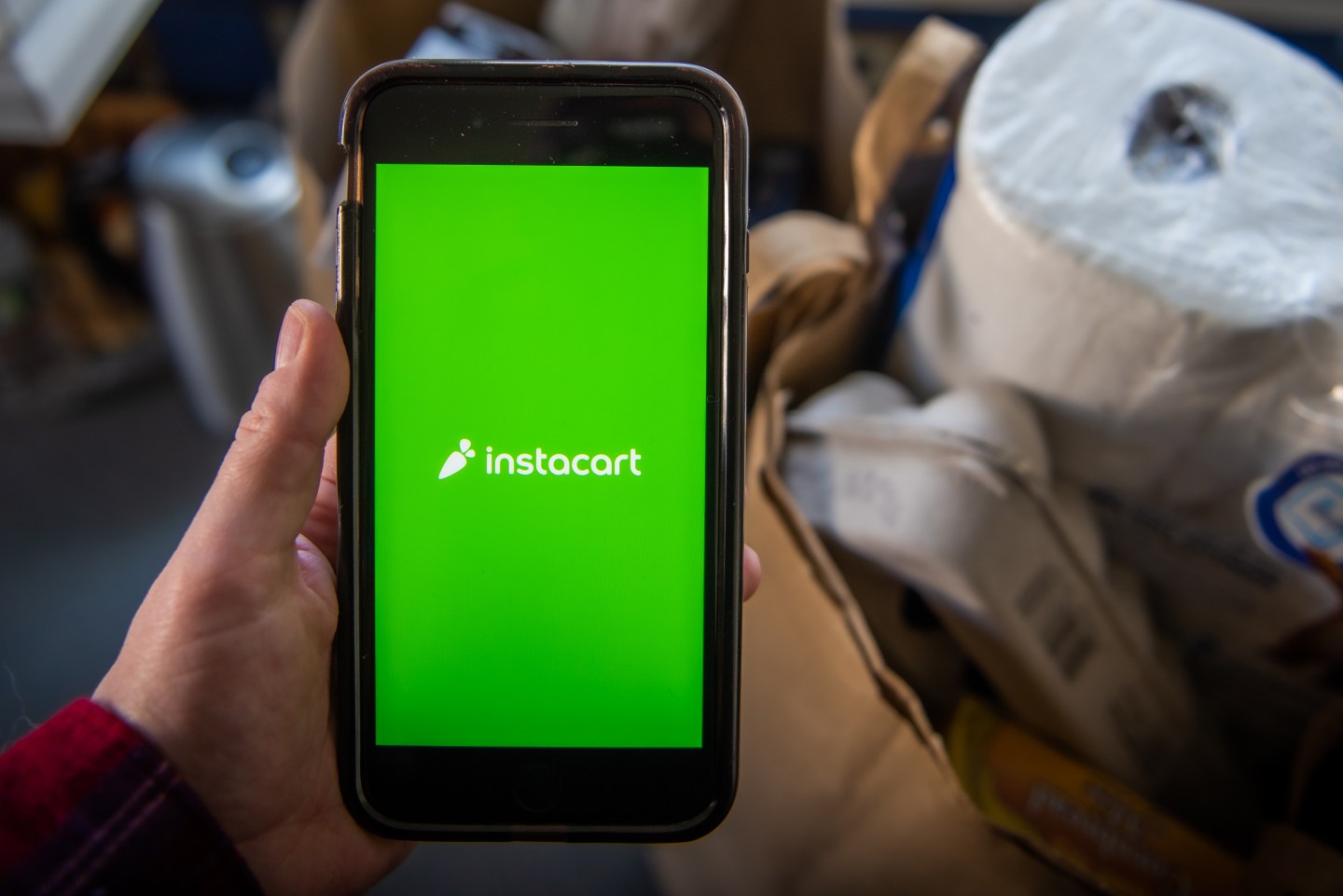 A person holding a cellphone with the Instacart logo on it, with a paper bag of groceries in the background