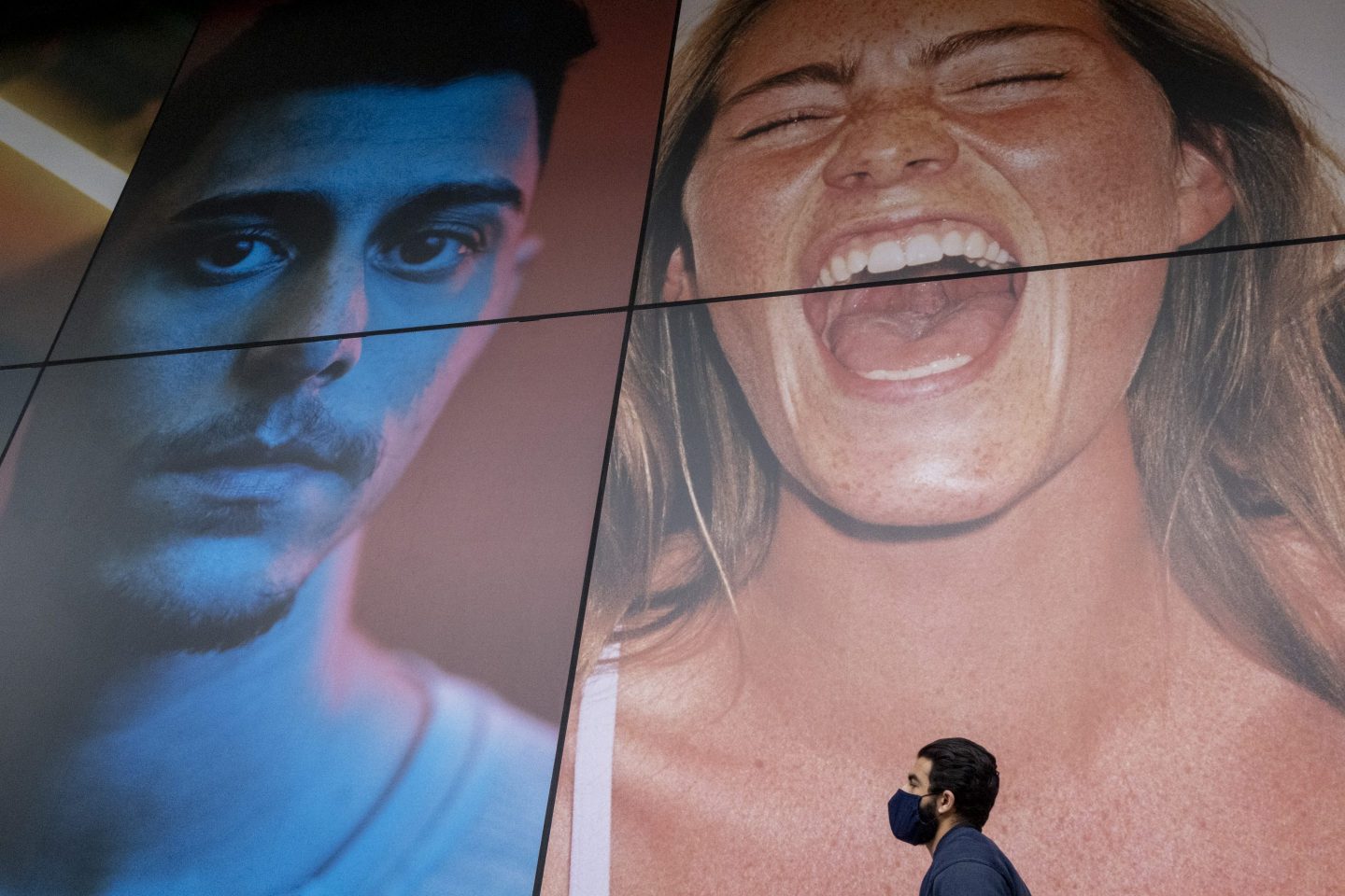 A man wearing a face covering walks beneath a large billboard featuring an ecstatic young woman
