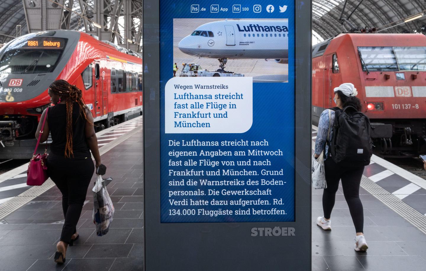 26 July 2022, Hessen, Frankfurt/Main: Rail passengers walk past an information screen in Frankfurt's main train station that reads "Lufthansa cancels almost all flights in Frankfurt and Munich" as a message.