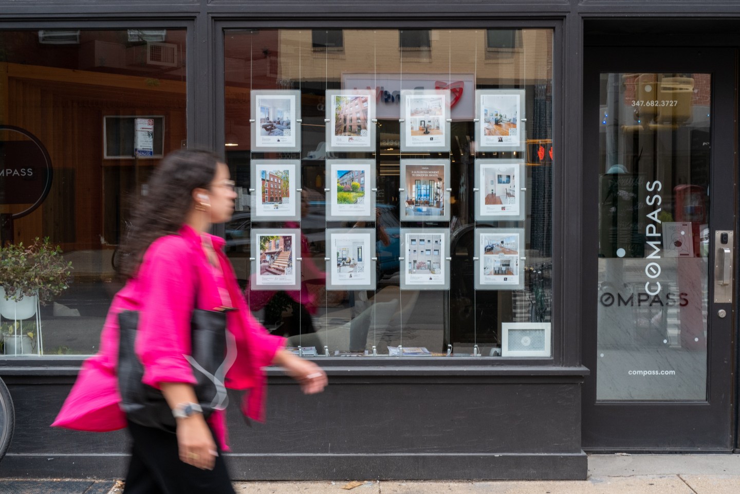 Woman walking in front of a real estate office in New York showing listings and prices.
