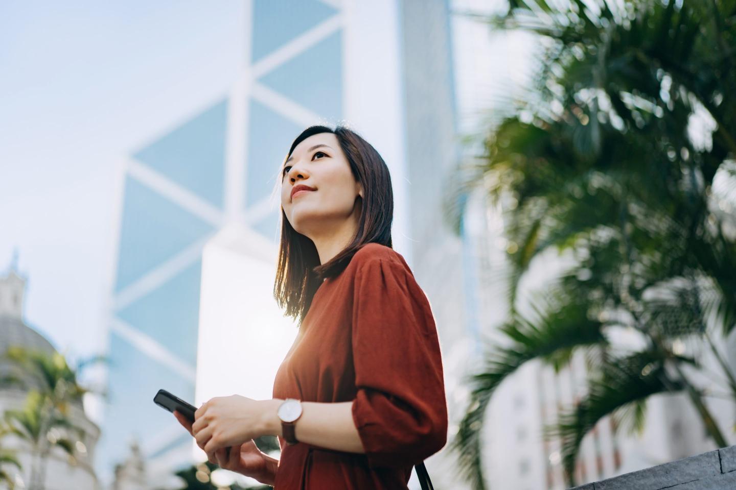 Confident and determined young Asian businesswoman looking away hopefully, using smartphone on the go against contemporary corporate skyscrapers in central business district in the city. Female leadership and determined to success