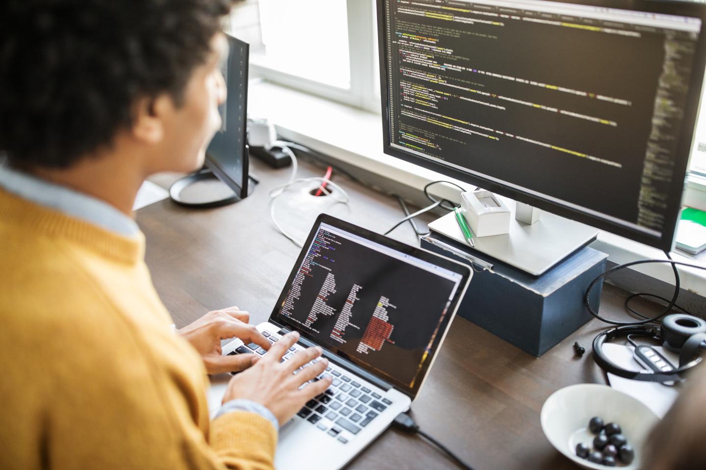 Businessman working on laptop and looking at desktop computer screen. Male professional sitting at his office desk and working on new software program.