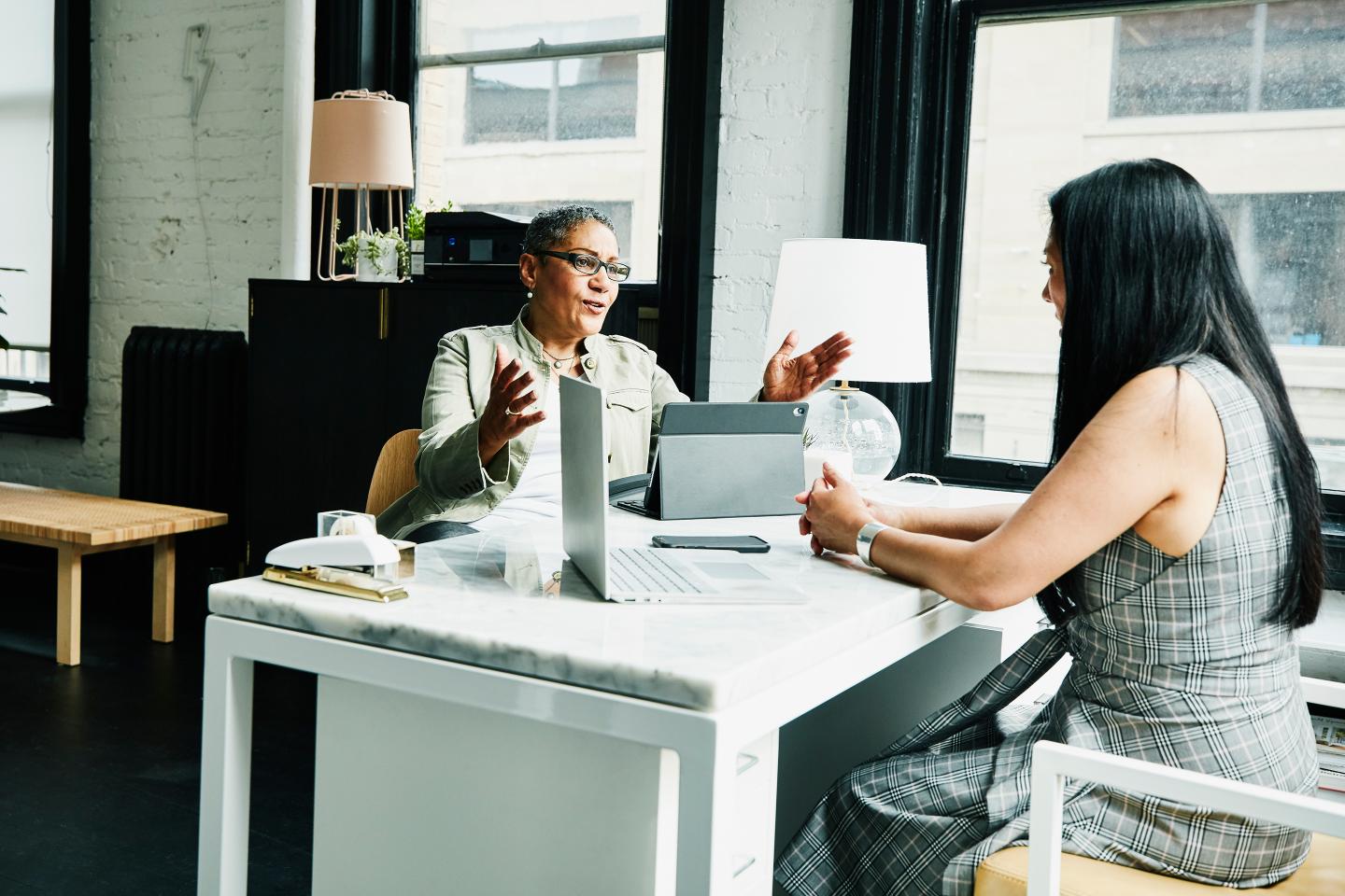 Woman of color speaking with another woman at a desk in office.