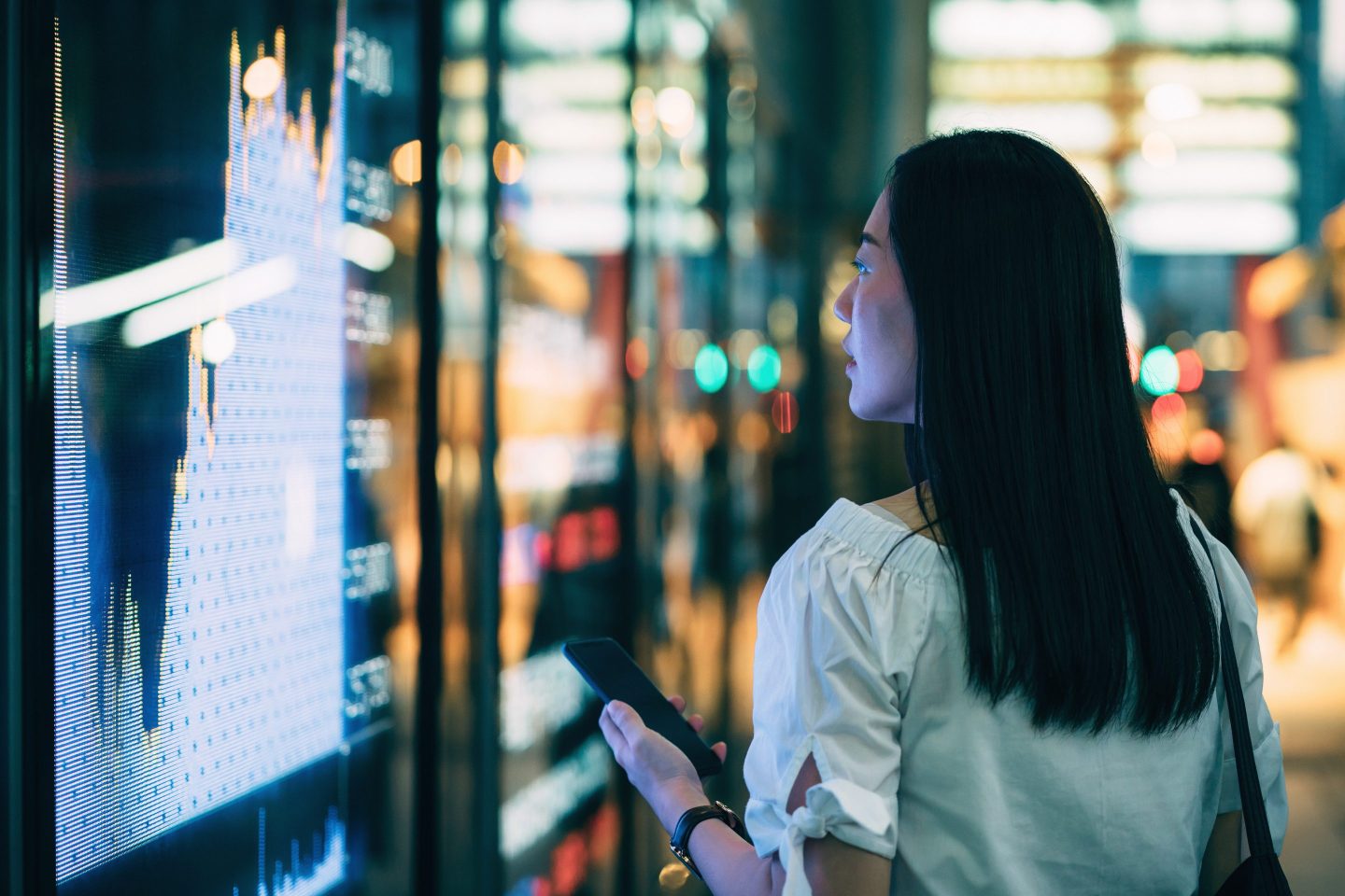 A young businesswoman checks financial trading data on smartphone by a stock market display