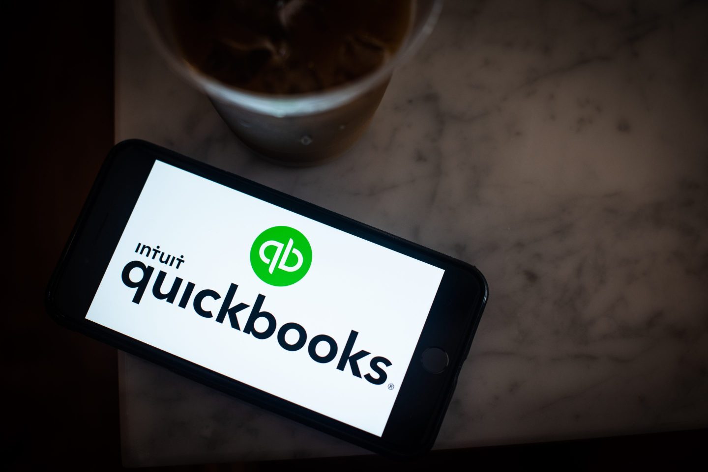 The Intuit Quickbooks logo on a cellphone next to a cup of iced coffee at a cafe