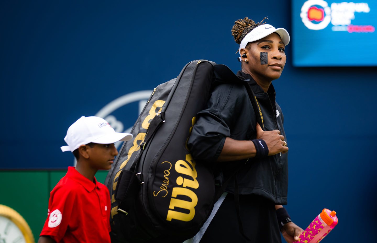 Serena Williams walking onto the court with her gear, with a young boy walking alongside her.