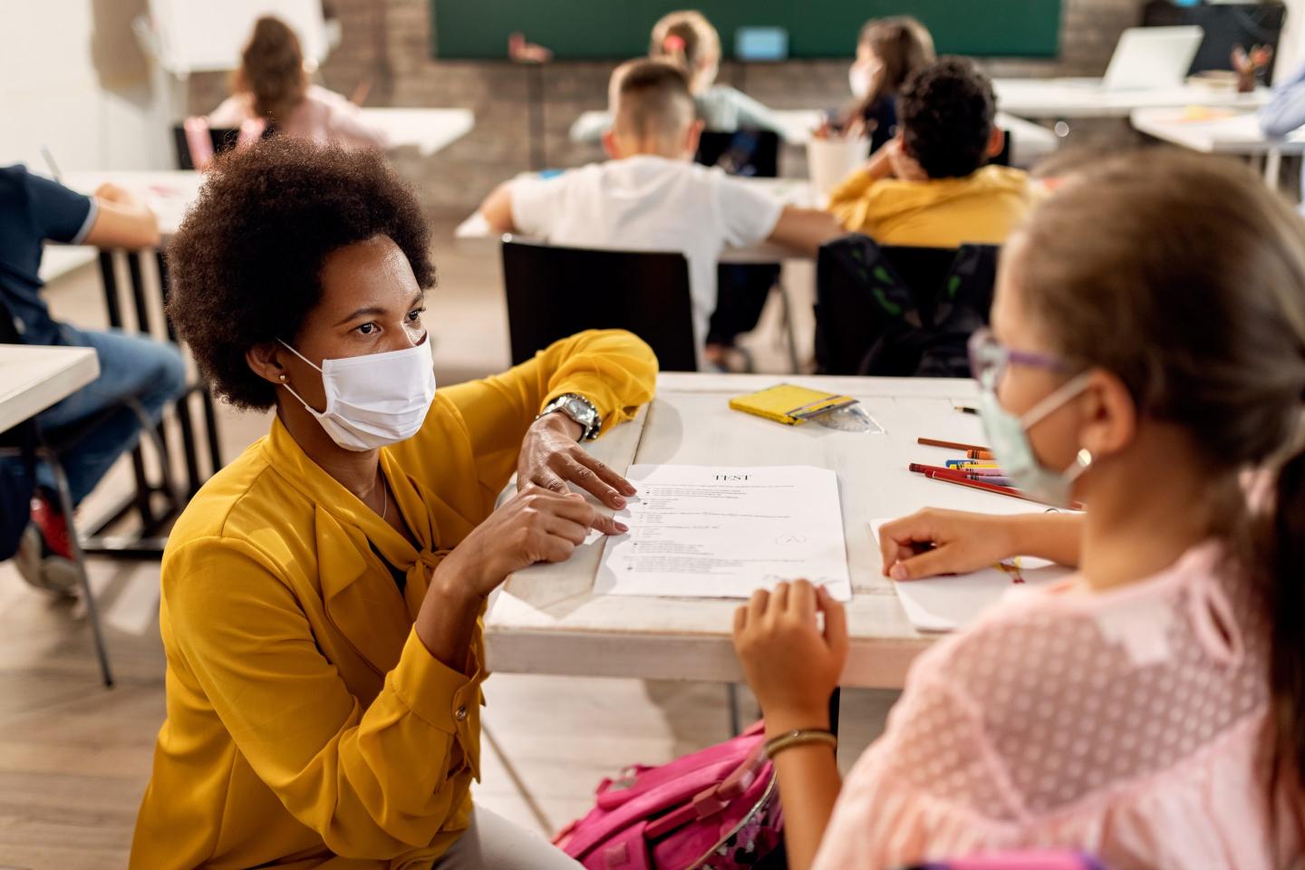 A Black teacher with a face mask explaining exam results to elementary student in the classroom.