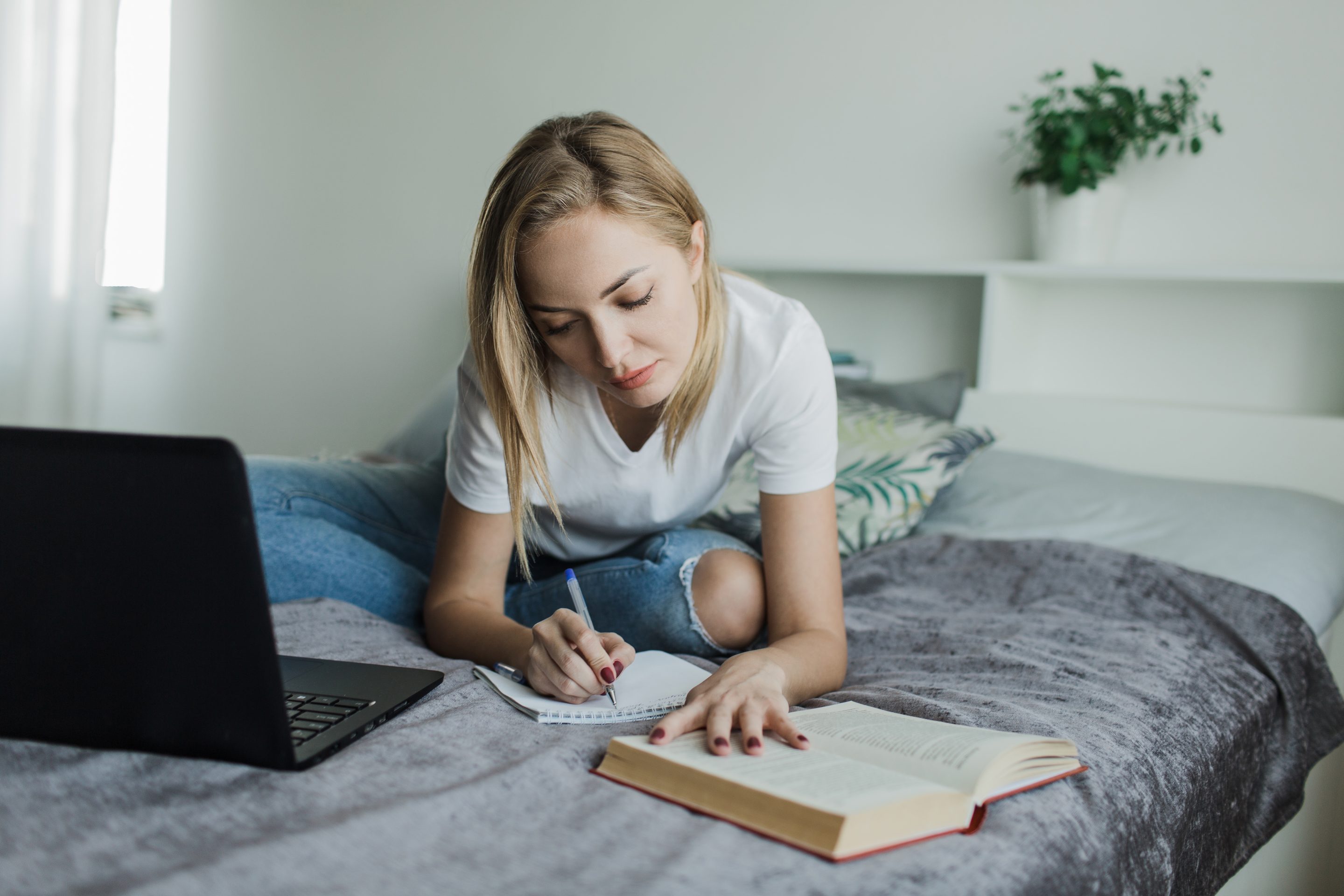 a student girl in a white t shirt and jeans is sitting on the bed studying writing reading