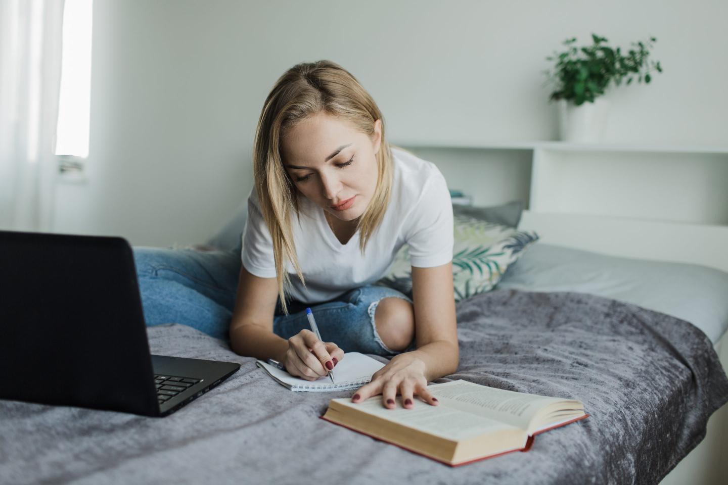a student girl in a white t shirt and jeans is sitting on the bed studying writing reading