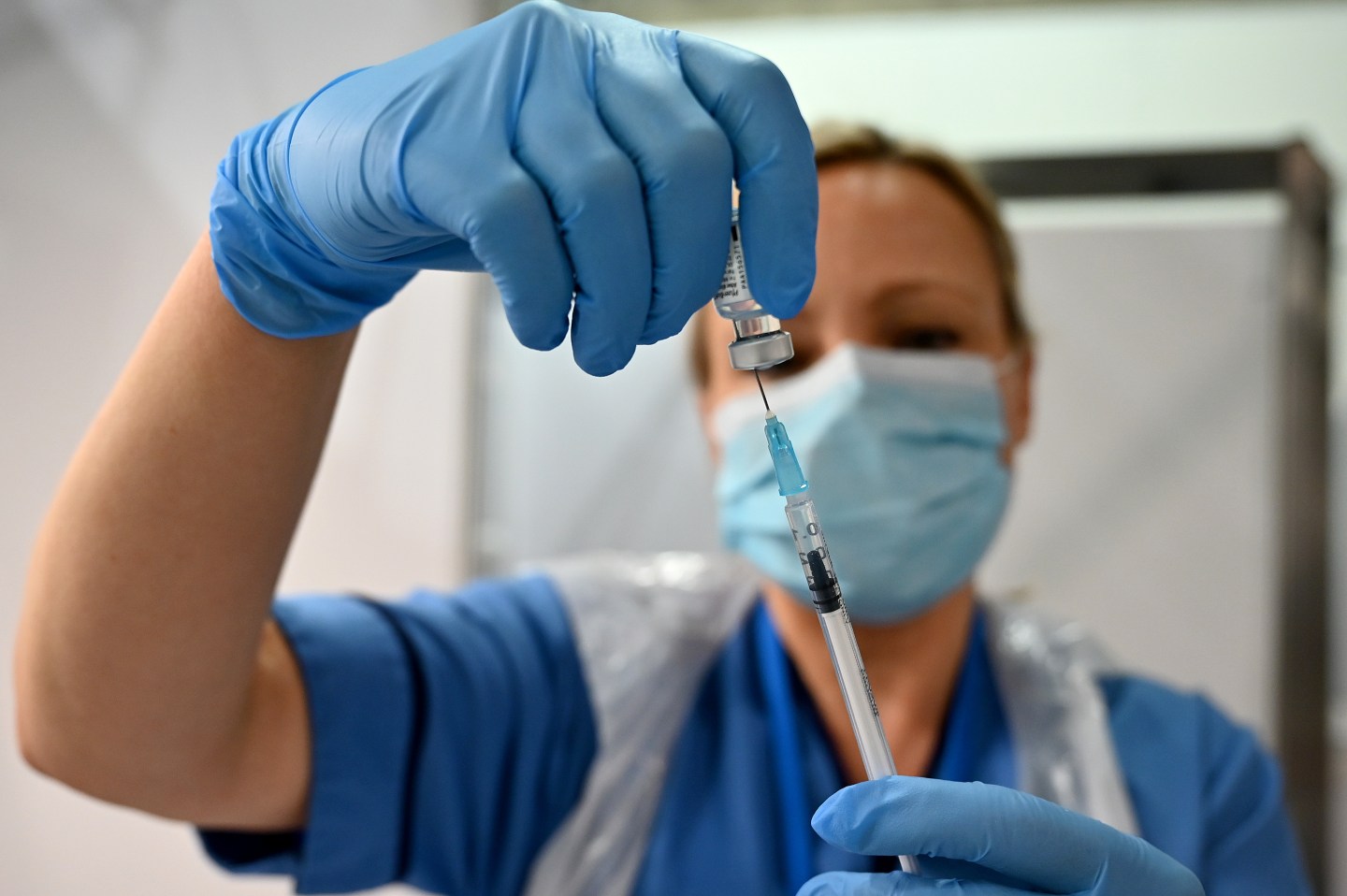 The image shows a British nurse in full PPE preparing a COVID vaccine.