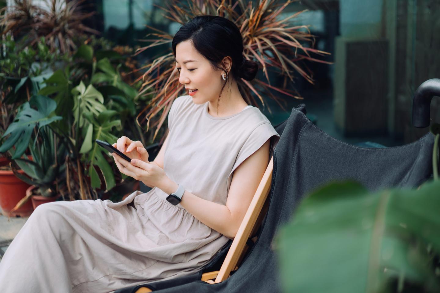 Young Asian woman using smartphone while relaxing on deck chair in the backyard, surrounded by beautiful houseplants