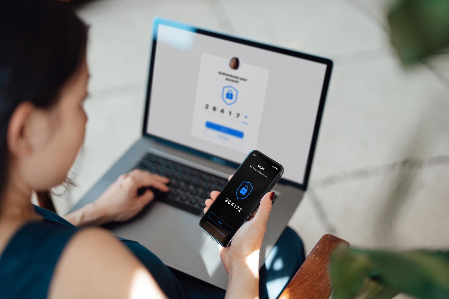 A woman holding her phone in her right hand with a laptop on her lap. A two-factor authentication screen is prompting her to sign in