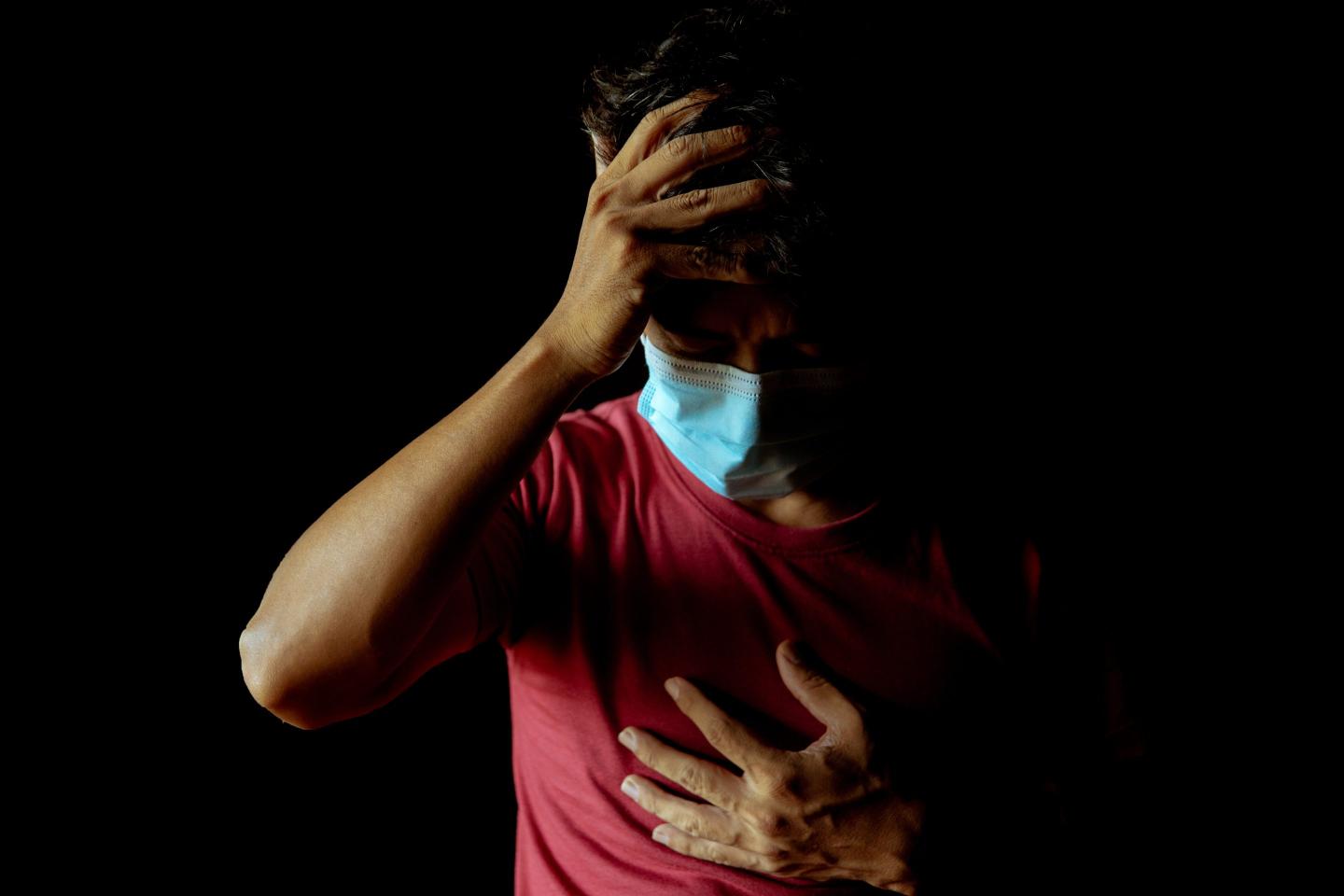 A long COVId patient clutches his chest and holds his head while wearing a surgical mask
