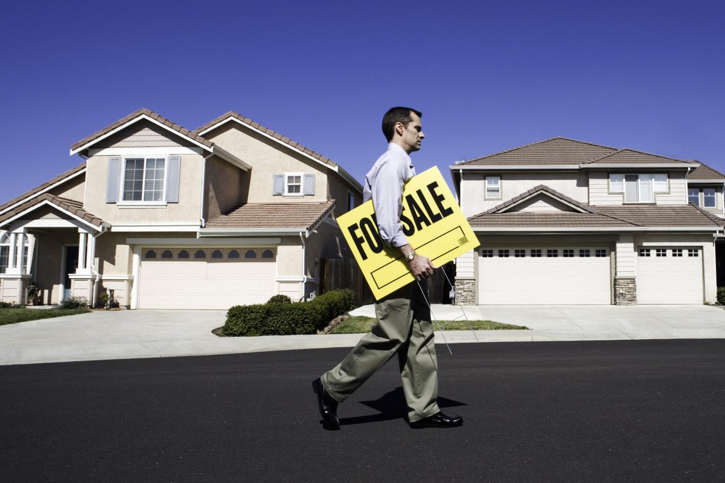 Male estate agent walking in front of house with holding for sale sign, side view