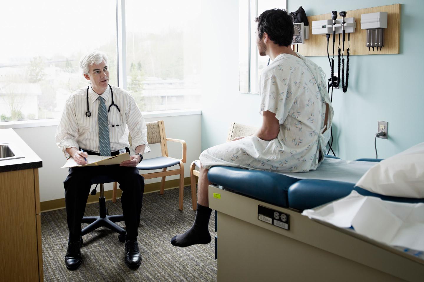 Male patient in gown sitting on exam table in discussion with doctor in exam room